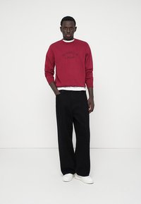 Röd sweatshirt med broderad text, kombinerad med svarta vida byxor och vita sneakers. Texturen är mjuk med en avslappnad passform.