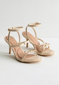 Beiga öppna tå high heel-sandaler med ankelremmar och dekorativa rosetter på tåremmarna, visade sida vid sida på en vit yta.