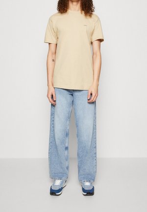 T-shirt en coton beige avec un col rond et un petit logo. Associé à un jean large bleu clair et des baskets de sport bleues.