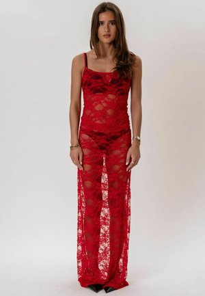 Femeie purtând o rochie lungă, roșie, din dantelă transparentă peste lenjerie neagră, stând cu brațele relaxate pe un fundal simplu, gri deschis.