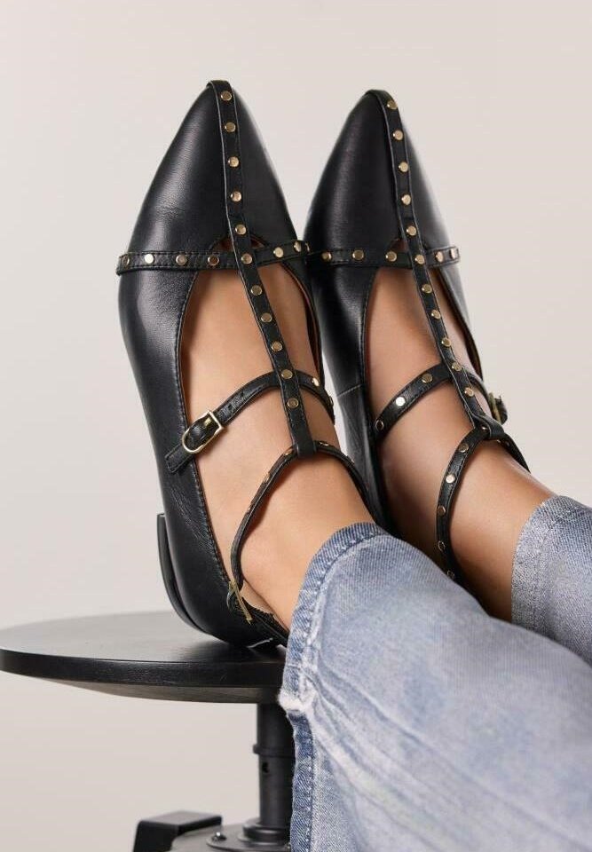Tacones de cuero negro con puntera afilada, que presentan correas y tachuelas doradas. Diseño estructurado con un tacón pequeño. Llevados con jeans claros.