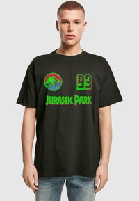 T-shirt en coton noir avec un graphique coloré d'un dinosaure et le texte "JURASSIC PARK 93" en vert et rose. Manches courtes, coupe décontractée.