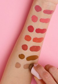 Campioni di vari colori per labbra applicati su un braccio, con tonalità di rosa, rosso e marrone in diverse texture lucide su uno sfondo rosa.