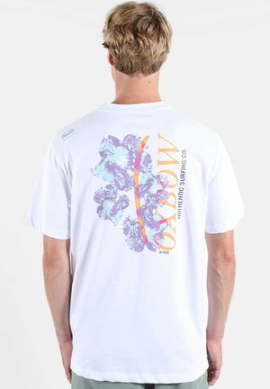 Personne blonde portant un T-shirt blanc avec des fleurs d'hibiscus violettes et bleues et un texte orange "Oxbow Authentic Surfing Co." au dos.