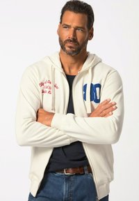 Bílá mikina na zip s kapucí, zdobená modro-červenými vyšívanými motivy, včetně čísla a textu. Noste přes tmavé tričko.