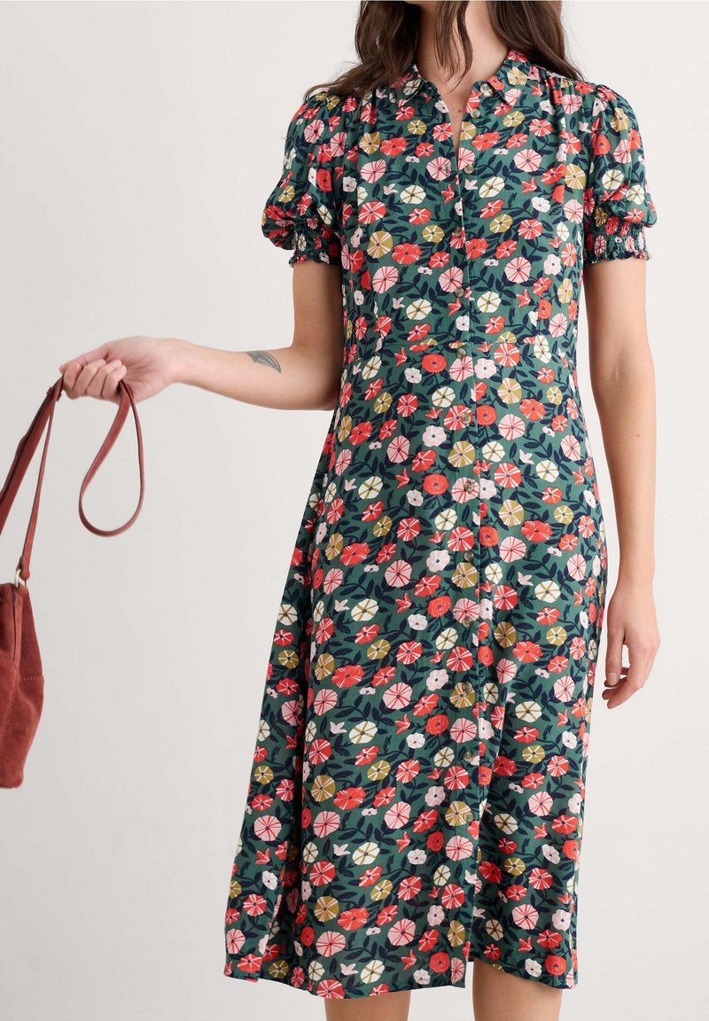 Femme portant une robe verte à fleurs avec des manches bouffantes tenant un sac à main marron par sa lanière à son côté.