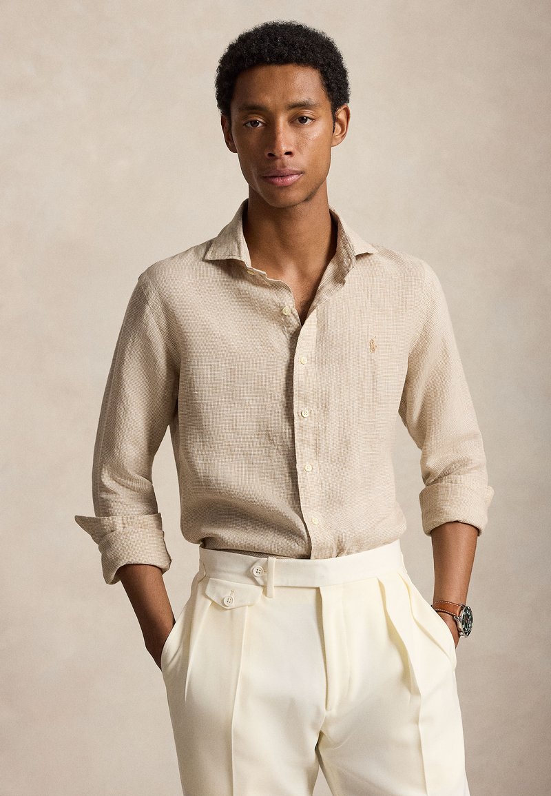 Uomo che indossa una camicia beige con texture e bottoni, maniche rimboccate e pantaloni bianchi plissettati a vita alta, mani nelle tasche, sfondo neutro.