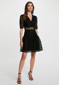 Abito in pizzo nero con scollo a V, maniche corte e vita aderente. Gonna svasata con orlo decorato e dettaglio di cintura a rete. Tacchi neri con cinturini.