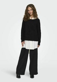 Black knitted sweater over a white button-up shirt, paired with wide black trousers and black ankle boots. Features long sleeves and textured fabric.