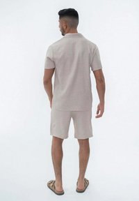 Polo shirt beige clair à manches courtes, associé à un short assorti. Texture lisse et design minimaliste. Des tongs marron complètent la tenue.