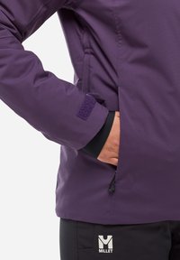 Person wearing a purple jacket with Velcro cuff, hand in side pocket, and black pants with a white Millet logo.