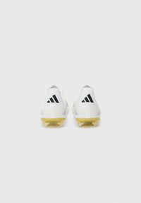 Des crampons de football blancs avec une tige texturée, une semelle extérieure dorée et trois rayures noires au niveau du talon. Conçus pour une meilleure traction sur les surfaces en herbe.