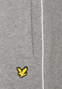 Grau meliertes Gewebe mit einer vertikalen Tasche und einem kleinen gelben Vogel-Logo in der Nähe der unteren linken Ecke neben einer weißen vertikalen Naht.