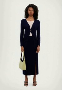 Cardigan azul-marinho sobre uma blusa creme, combinado com uma saia azul-marinho com uma fenda lateral. Segurando uma carteira verde-clara, usando sandálias pretas.