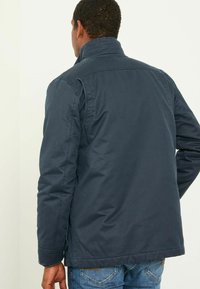 Marineblaue Jacke aus leichtem Material mit hohem Kragen, sichtbaren Nähten und geradem Schnitt. Die Rückansicht zeigt eine strukturierte Oberfläche.