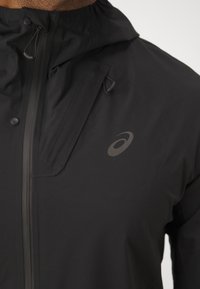 Chaqueta deportiva negra con cierre frontal de cremallera, capucha ajustable y detalle de logo. Fabricada con un tejido ligero y resistente a las inclemencias del tiempo.