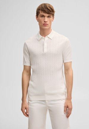 Giovane uomo con capelli corti castani che indossa una polo bianca a maniche corte con motivo a trecce e pantaloni bianchi, in piedi davanti a uno sfondo semplice.