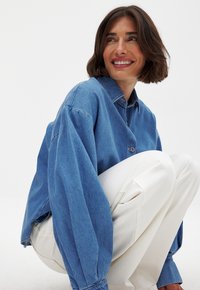 Denim shirt in light blue with a collared neck, button front, and long, loose sleeves, paired with white textured pants.