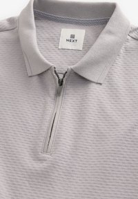 Hellgraues Poloshirt mit Reißverschlusskragen. Strukturiertes Gewebe mit einem subtilen Muster und einem kontrastierenden Karomuster am inneren Kragen. Markenlabel sichtbar.