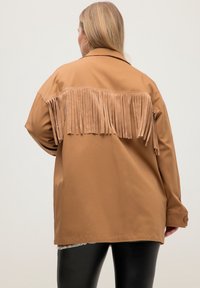 Giacca marrone con una texture liscia, caratterizzata da un dettaglio con frange sulla schiena. Vestibilità comoda con colletto e polsini abbottonati.
