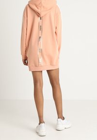 Mikina s kapucí v měkké broskvové tkanině, s bílými bočními panely a reflexním logem na zadní straně. Žebrovaný lem a manžety.