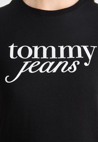 Svart t-skjorte med fet, hvit "tommy jeans" grafisk tekst. Stoffet er glatt med en klassisk rund halsdesign og korte ermer.