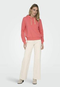 Sudadera de color coral con cordones dorados y un pequeño detalle de corazón, combinada con pantalones de pierna ancha en color blanco roto y zapatos de color beige claro. Textura de tela suave.