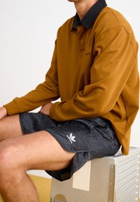 Brun långärmad polo skjorta med svart krage, tillsammans med svarta mönstrade shorts med vit Adidas-logotyp, sittande på en metallisk yta.