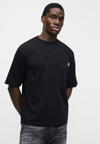 Μαύρο βαμβακερό μπλουζάκι με στρογγυλό λαιμό, κοντά μανίκια και τσέπη στο μπροστινό μέρος με κεντημένες λεπτομέρειες. Φοριέται με γκρι τζιν με φθορές.