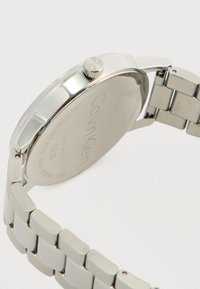 Ρολόι από ανοξείδωτο ατσάλι με στρογγυλή θήκη, φινίρισμα ασημί, υφασματοποιημένο κορώνα και λουράκι τύπου λινκ; χαραγμένο λογότυπο της μάρκας στην πίσω πλευρά.