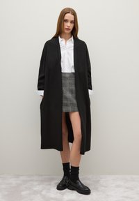 Cardigan lungo nero con apertura frontale, texture morbida, abbinato a una camicia bianca con bottoni, gonna mini a quadri, calze nere e scarpe chunky.