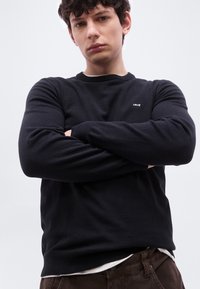 Jeune homme portant un sweat-shirt noir Levi's et un pantalon marron, debout les bras croisés devant un fond clair uni.