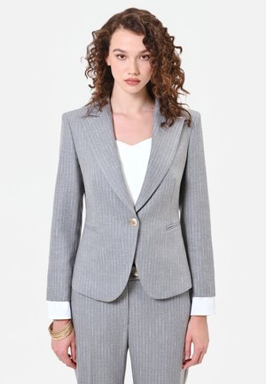 Donna con capelli ricci che indossa una giacca di abito a righe grigia e pantaloni abbinati, con polsini bianchi della camicia visibili e braccialetti dorati al polso sinistro.
