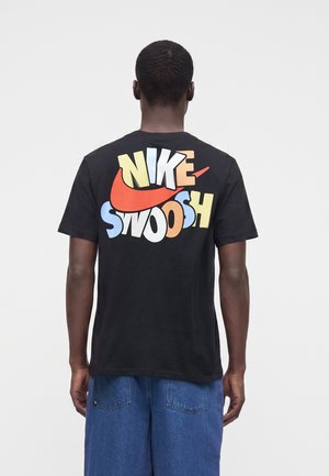 Άτομο που φοράει μαύρο T-shirt με πολύχρωμο κείμενο "NIKE SWOOSH" και κόκκινο λογότυπο swoosh στην πλάτη, συνδυασμένο με μπλε τζιν παντελόνια, με την πλάτη προς τα εμπρός.