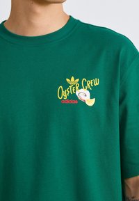 Grön bomullströja med korta ärmar, med en gul och röd "Oyster Crew" grafik och en Adidas-logotyp. Enkel krage i crew neck-design.