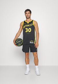 Camiseta de baloncesto negra con acentos amarillos y "SAN FRANCISCO 30" impreso. El modelo sostiene un balón de baloncesto negro y verde, vistiendo pantalones cortos negros y zapatillas blancas.
