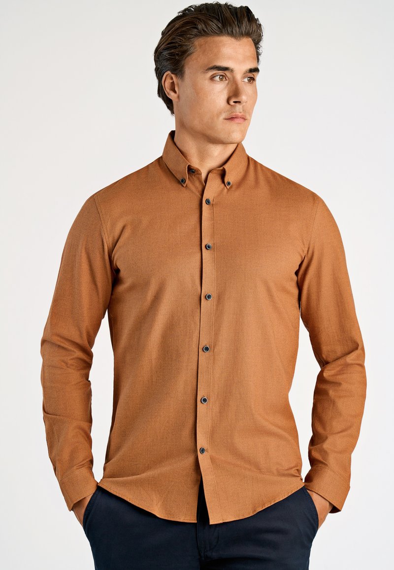 Jasnobrązowa koszula zapinana na guziki z fakturowanej tkaniny, czarnymi guzikami, dopasowanym krojem i długimi rękawami, w połączeniu z ciemnymi spodniami.