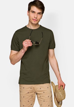 Oliwkowa bawełniana koszulka z krótkim rękawem, okrągłym dekoltem i małym logo. Model trzyma okulary przeciwsłoneczne, założone do wzorzystych beżowych szortów i słomkowego kapelusza.