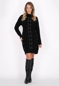 Abito in maglia nera con collo alto, caratterizzato da una vestibilità dritta, decorazioni in perle e maniche lunghe. Abbinato a stivali neri al ginocchio.