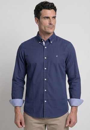 Hombre con camisa azul marino abotonada y mangas remangadas, y pantalones beige, mirando ligeramente hacia la derecha sobre un fondo liso.