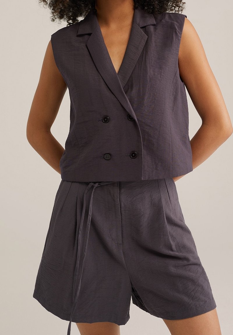 Grå linne ärmlös kavaj med dubbelknäppning och två svarta knappar samt matchande plisserade shorts med dragsko i midjan, slät textur.