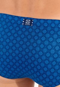 Blå mønstrede badebukser med diamantdesign, blød tekstur og et lille label med teksten "H BEACHWEAR" nær taljen.