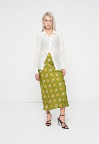 Camisa branca transparente abotoada por cima de uma saia maxi verde com padrões que apresentam motivos de olhos e designs florais. Sandálias de salto altas pretas.
