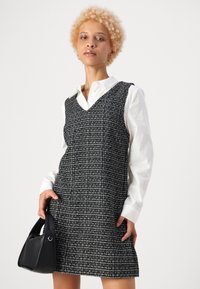 Črno-bela tweed obleka z V-izrezom, brez rokavov in oprijetim krojem, v kombinaciji z belo s srajco in črnim ročnim žepkom.