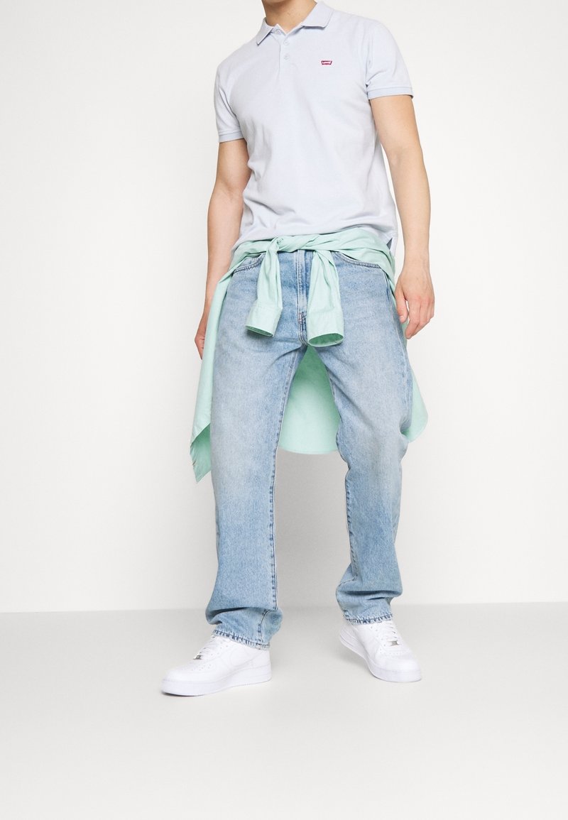 Homme portant un polo bleu clair, un jean clair, des baskets blanches, avec une chemise vert clair nouée autour de la taille, sur fond blanc uni.