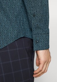 Shirt van donker teal stof met een herhalend lichtblauw "HUGO" patroon, met knoopmanchetten, gecombineerd met donker geruite broek.