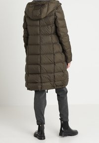 Lungo cappotto imbottito verde oliva con design trapuntato, cappuccio e cordoncino elastico sull'orlo. Abbinato a pantaloni grigi e stivaletti neri.