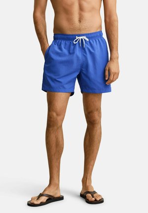 Mann trägt blaue Badeshorts mit weißem Kordelzug und braunen Flip-Flops, steht mit einer Hand in der Tasche vor einfachem Hintergrund.