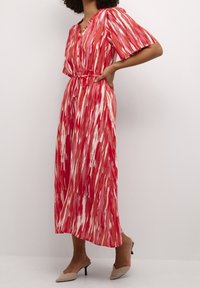 Robe longue rayée rouge et blanche avec un col en V, des manches courtes, une ceinture à nouer à la taille et une silhouette fluide. Portée avec des talons beige.