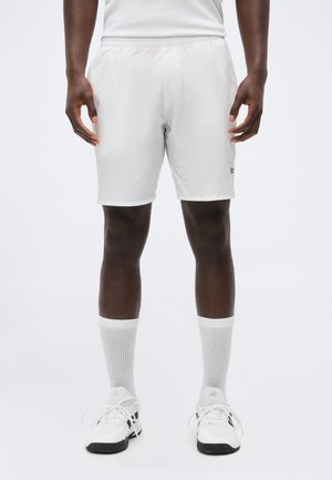 Mężczyzna ubrany w białe sportowe spodenki, białe skarpetki do kostek i białe tenisówki Adidas stoi na jednolitym tle.